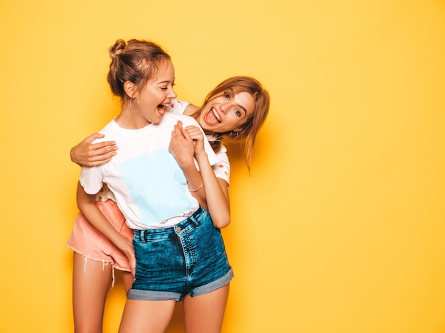 Twee jonge mooie glimlachende hipster meisjes in trendy zomerkleren. Sexy onbezorgde vrouwen die dichtbij gele muur stellen. Positieve modellen worden gek en hebben plezier