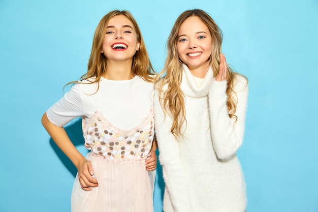 Twee jonge mooie glimlachende hipster vrouwen in trendy zomer witte kleding. Sexy onbezorgde vrouwen die dichtbij blauwe muur stellen. Positieve modellen