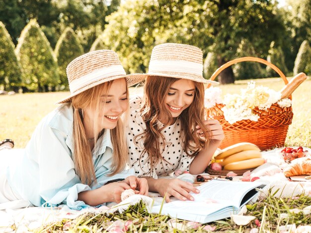 Twee jonge mooie glimlachende vrouw in trendy zomerjurk en hoeden. Zorgeloze vrouwen die buiten picknicken.