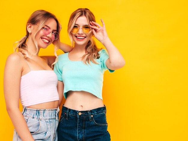 Twee jonge mooie lachende blonde hipster vrouw in trendy zomerkleren Sexy zorgeloze vrouwen poseren in de buurt van gele muur in studio Positieve modellen plezier vrolijk en gelukkig In zonnebril