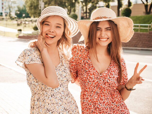 Twee jonge mooie lachende hipster meisjes in trendy zomer sundress. Sexy zorgeloze vrouwen die zich voordeed op de straat achtergrond in hoeden. Positieve modellen die plezier hebben en knuffelen.Ze vertonen vredesteken en tong