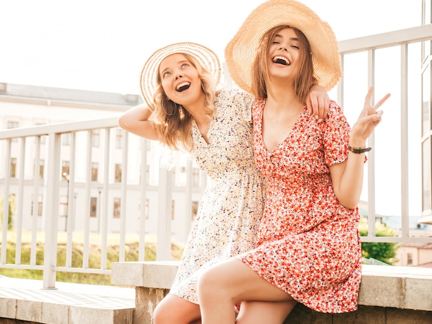 Twee jonge mooie lachende hipster meisjes in trendy zomer sundress. Sexy zorgeloze vrouwen die zich voordeed op de straat achtergrond in hoeden. Positieve modellen die plezier hebben en knuffelen. Ze vertonen vredesteken