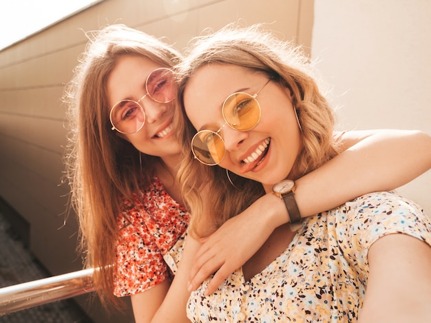 Twee jonge mooie lachende hipster meisjes in trendy zomer sundress. sexy zorgeloze vrouwen die zich voordeed op de straat achtergrond in zonnebril. ze nemen selfie-zelfportretfoto's op smartphone bij zonsondergang