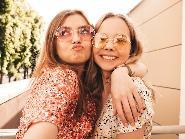 Twee jonge mooie lachende hipster meisjes in trendy zomer sundress. sexy zorgeloze vrouwen die zich voordeed op de straat achtergrond in zonnebril. ze nemen selfie-zelfportretfoto's op smartphone bij zonsondergang