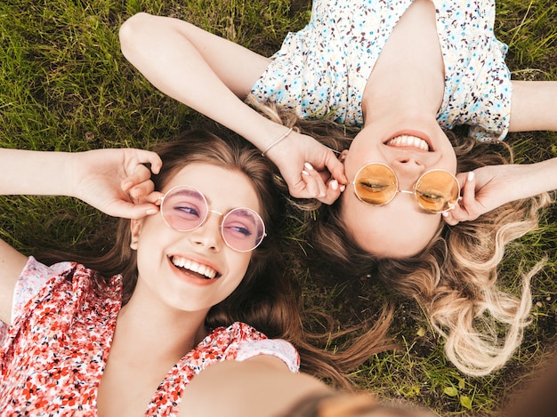 Twee jonge mooie lachende hipster meisjes in trendy zomer zonnejurk. sexy zorgeloze vrouwen liggend op het groene gras in zonnebril. positieve modellen plezier. bovenaanzicht. selfie foto's maken op smartphone