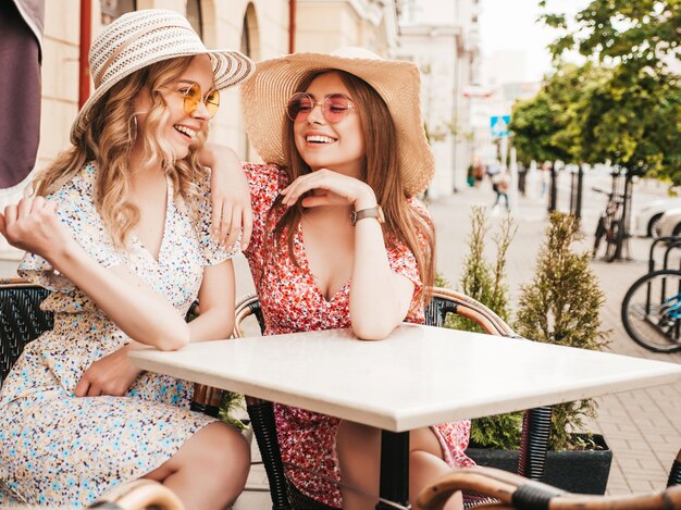 Twee jonge mooie lachende hipster meisjes in trendy zomer zonnejurk. Zorgeloze vrouwen chatten in veranda cafe op de straat achtergrond in zonnebril. Positieve modellen met plezier en communiceren