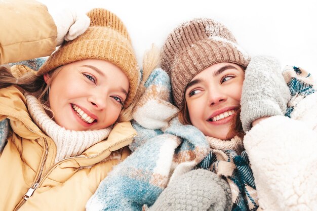 Twee jonge mooie lachende hipster vrouw in trendy warme kleding en sjaals. Zorgeloos vrouwen poseren in de straat in het park. Positieve pure modellen die in de sneeuw liggen. Genieten van winterse momenten. kerst concept