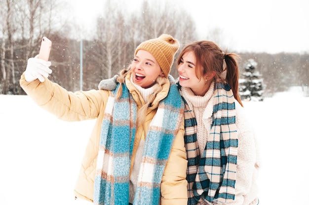 Twee jonge mooie lachende hipster vrouw in trendy warme kleding en sjaals. zorgeloos vrouwen poseren in de straat in het park. positieve pure modellen die plezier hebben in de sneeuw. genieten van winterse momenten. selfie maken