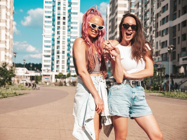 Twee jonge mooie lachende hipster vrouw in trendy zomerkleren. Sexy zorgeloze vrouwen poseren in de straat met roze haren. Positieve pure modellen die plezier hebben bij zonsondergang. Vrolijk en gelukkig