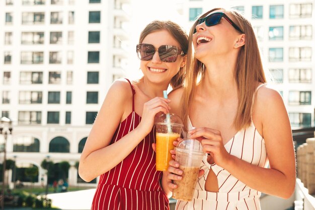 Twee jonge mooie lachende hipster vrouw in trendy zomerkleren Zorgeloze vrouwen poseren buitenshuis Positieve modellen houden en drinken van verse cocktail smoothie drankje in plastic beker met stro