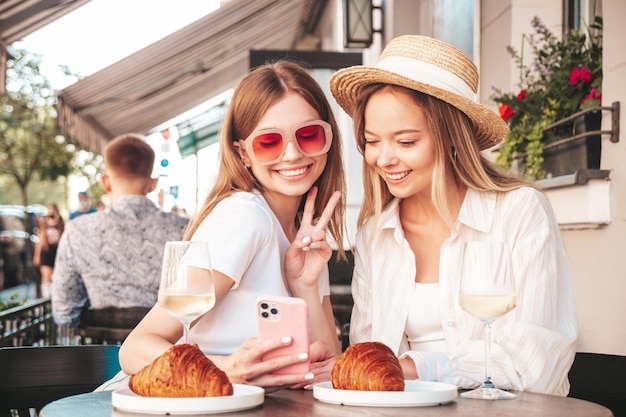 Twee jonge mooie lachende hipster vrouw in trendy zomerkleren zorgeloze vrouwen poseren op veranda café in de straat positieve modellen die witte wijn drinken croissant eten selfie foto's maken