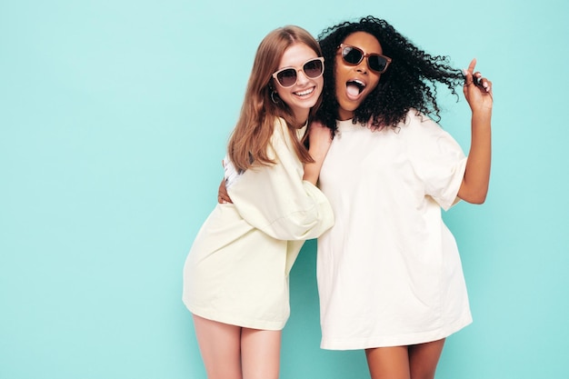 Twee jonge mooie lachende internationale hipster vrouw in trendy zomerkleren Sexy zorgeloze vrouwen poseren in de buurt van blauwe muur in studio Positieve modellen plezier in zonnebril Concept van vriendschap