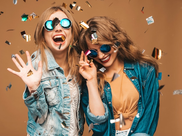 Twee jonge mooie lachende meisjes in trendy zomer kleding en zonnebril. Sexy zorgeloze vrouwen poseren. Positieve schreeuwende modellen onder confetti