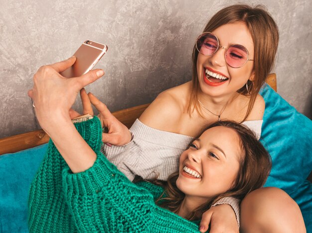 Twee jonge mooie lachende prachtige meisjes in trendy zomerkleren. Sexy zorgeloze vrouwen poseren in interieur en nemen selfie. Positieve modellen met plezier met smartphone.