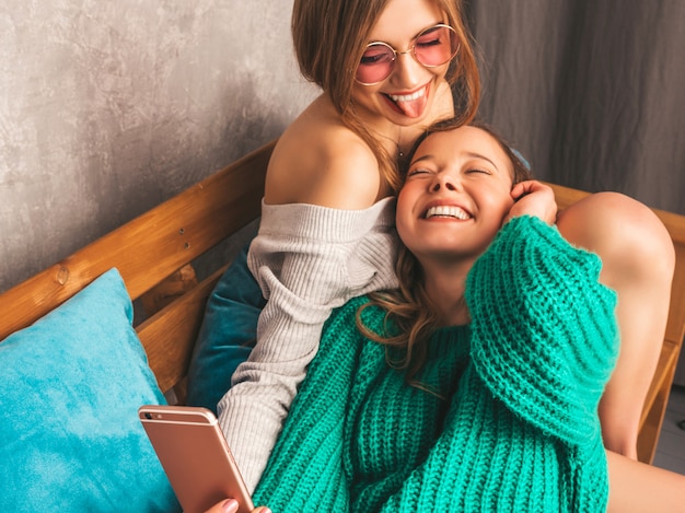 Twee jonge mooie lachende prachtige meisjes in trendy zomerkleren. Sexy zorgeloze vrouwen poseren in interieur en nemen selfie. Positieve modellen met plezier met smartphone.