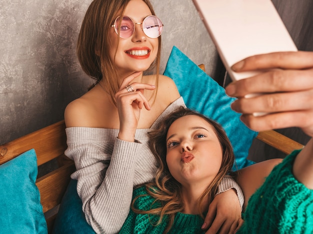 Twee jonge mooie lachende prachtige meisjes in trendy zomerkleren. Sexy zorgeloze vrouwen poseren in interieur en nemen selfie. Positieve modellen met plezier met smartphone.