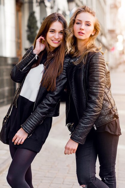 Twee jonge mooie meisjes poseren buitenshuis samen. Lifestyle stedelijke sfeer. Centrum stad achtergrond. Beste vrienden die zwarte casual herfstoutfit dragen.