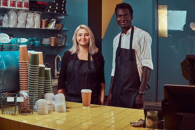 Twee jonge multiraciale barista's in schorten staan gastvrij in hun trendy coffeeshop.