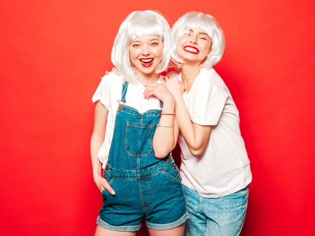 Twee jonge sexy lachende hipster meisjes in witte pruiken en rode lippen. Mooie trendy vrouwen in de zomer kleding. Zorgeloze modellen poseren in de buurt van rode muur in studio zomer gek