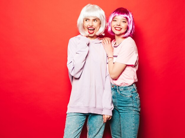 Twee jonge sexy lachende hipster meisjes in witte pruiken en rode lippen. Mooie trendy vrouwen in zomer kleding. Zorgeloze modellen poseren in de buurt van rode muur in studio gek