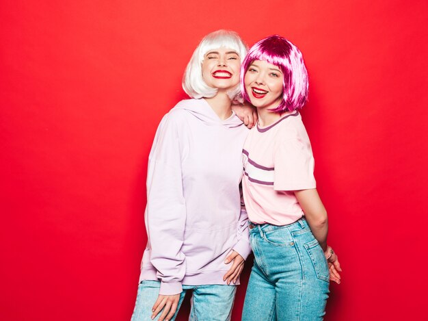 Twee jonge sexy lachende hipster meisjes in witte pruiken en rode lippen. Mooie trendy vrouwen in zomer kleding. Zorgeloze modellen poseren in de buurt van rode muur in studio gek