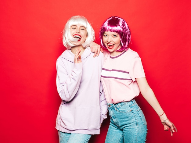 Twee jonge sexy lachende hipster meisjes in witte pruiken en rode lippen. Mooie trendy vrouwen in zomer kleding. Zorgeloze modellen poseren in de buurt van rode muur in studio gek