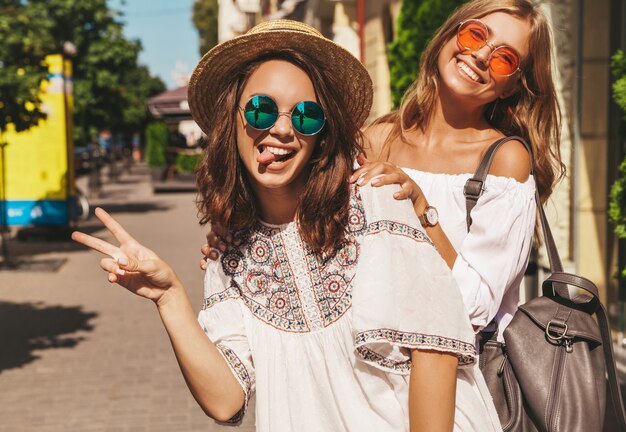 Twee jonge stijlvolle hippie brunette en blonde vrouwen modellen in zonnige zomerdag in witte hipster kleding poseren