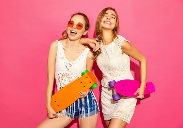 Twee jonge stijlvolle lachende blonde vrouwen met penny skateboards. Modellen in de zomer hipster witte kleren die dichtbij roze muur in zonnebril stellen. Positieve vrouw