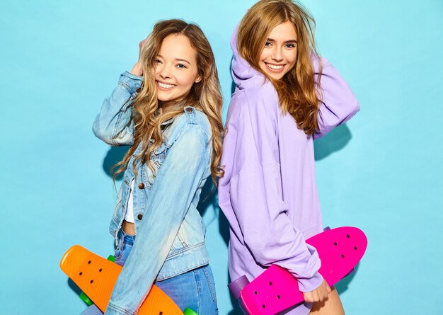 Twee jonge stijlvolle lachende blonde vrouwen met penny skateboards. Modellen in zomer hipster sport kleding poseren in de buurt van blauwe muur. Positieve vrouw