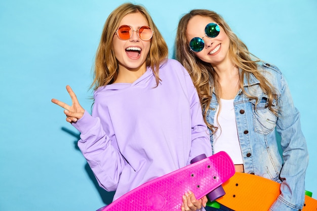 Twee jonge stijlvolle lachende blonde vrouwen met penny skateboards. Modellen in zomer hipster sport kleding poseren in de buurt van blauwe muur. Positieve vrouwen worden gek