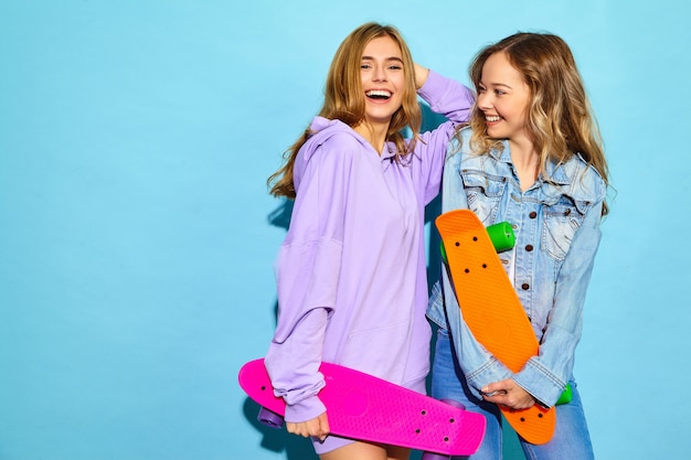 Twee jonge stijlvolle lachende blonde vrouwen met penny skateboards. Modellen in zomer hipster sport kleding poseren in de buurt van blauwe muur. Positieve vrouwen worden gek