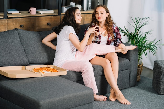 Twee jonge vrouwen die op bank roosterende glazen zitten