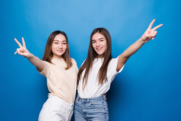 Twee jonge vrouwen die vingers tonen die overwinningsteken doen dat over blauwe muur wordt geïsoleerd. Nummer twee.