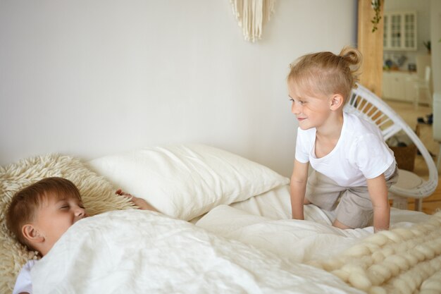 Twee jongens spelen in bed. Schattige blonde kleine jongen zittend op witte bedkleren kijken naar zijn bejaarde broer die doet alsof hij slaapt. Kinderen spelen in de slaapkamer. Familie, jeugd en plezier