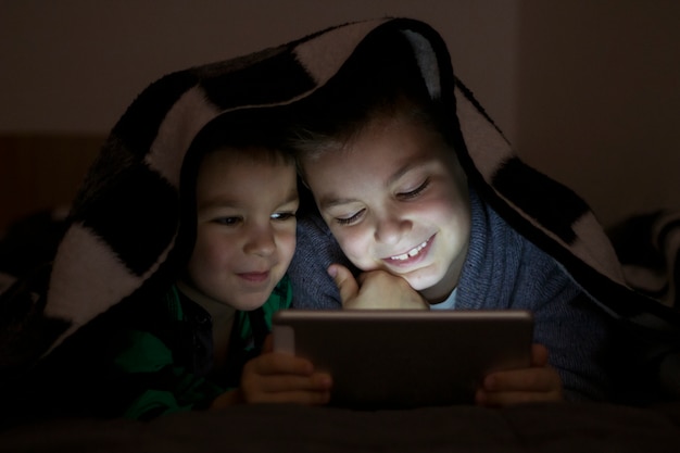 Twee kinderen met behulp van tablet pc onder de deken &#39;s nachts. Leuke Broers Met Tablet Computer In Een Donkere Kamer Lachend.