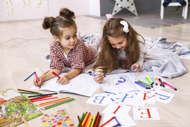 Twee kleine blije meisjes die tekenen in het kleurboek op de vloer op de deken liggen en letters leren