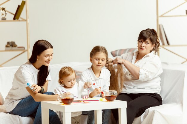 Twee kleine meisjes hun aantrekkelijke jonge moeder en hun charmante grootmoeder zittend op de bank en tijd samen thuis doorbrengen. Generatie van vrouwen. Internationale Vrouwendag. Gelukkige Moederdag.