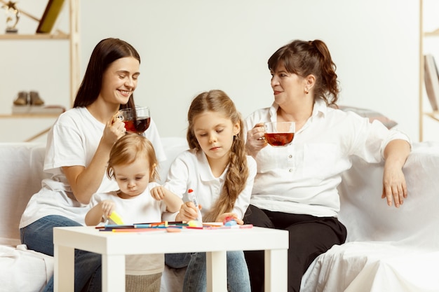 Twee kleine meisjes hun aantrekkelijke jonge moeder en hun charmante grootmoeder zittend op de bank en tijd samen thuis doorbrengen. Generatie van vrouwen. Internationale Vrouwendag. Gelukkige Moederdag.