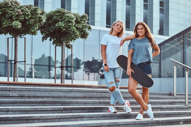 Twee lachende jonge meisjes houden een skateboard vast terwijl ze op trappen staan op de achtergrond van de wolkenkrabber.