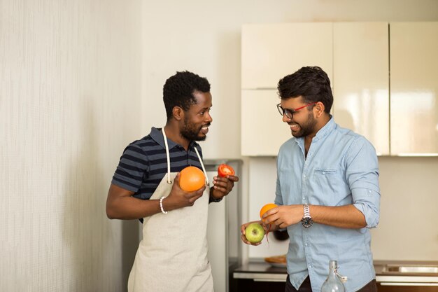 Twee mannelijke studenten in vrijetijdskleding gaan smoothie koken in de keuken van het hostel, grapefruit, sinaasappel, appel, tomaat in handen houdend, vrolijk pratend en lachend.