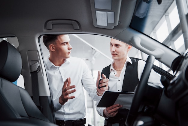 Twee mannen staan in de showroom tegen auto's. Close-up van een sales manager in een pak dat een auto aan een klant verkoopt. De verkoper geeft de sleutel aan de klant.