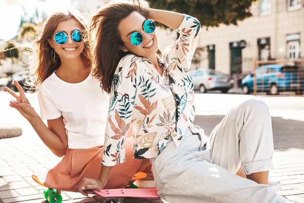 Twee modellen in zonnige zomerdag in hipster kleding zittend op cent skateboard op straat