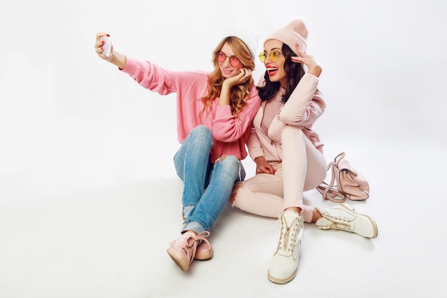 Twee modieuze meiden die zelfportret maken. Witte achtergrond. Roze kleren.