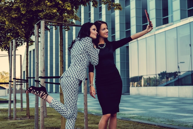 Twee modieuze vrouwen in stijlvolle kleding en zonnebril poseren midden in het zakelijke stadsdistrict.