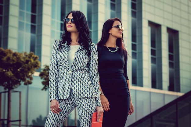 Twee modieuze vrouwen in stijlvolle kleding en zonnebril poseren midden in het zakelijke stadsdistrict.