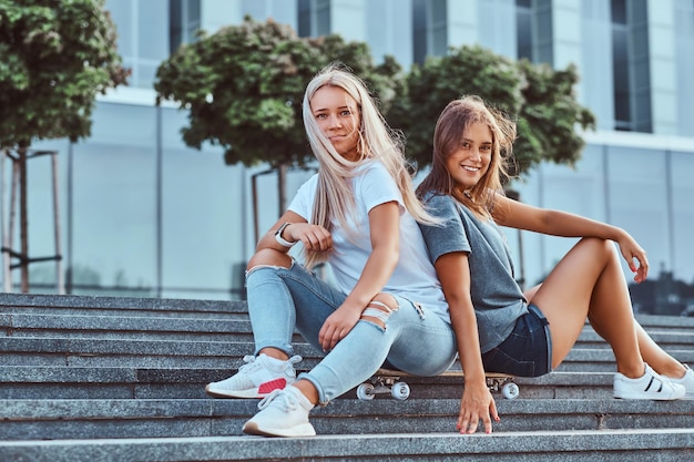 Twee mooie hipstermeisjes die op stappen zitten met een skateboard op een achtergrond van de wolkenkrabber.