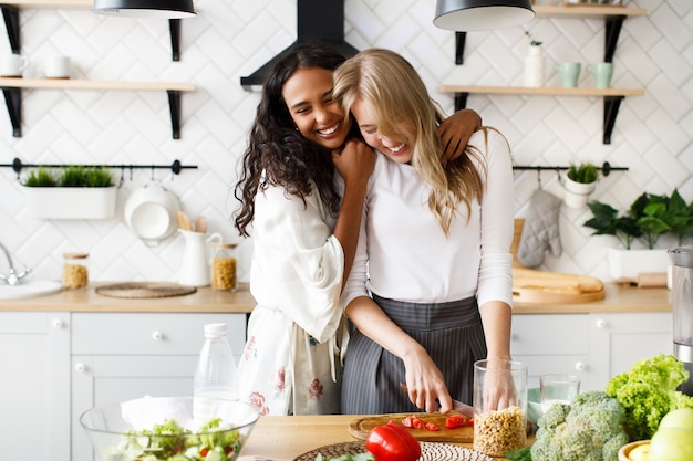 Twee mooie jonge vrouwen maken een gezond ontbijt en knuffelen in de buurt van de tafel vol met verse groenten op de witte moderne keuken