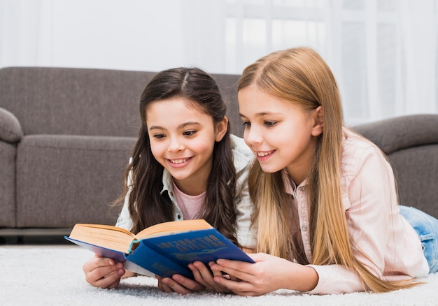 Twee mooie meisjes die op tapijt liggen die het boek samen thuis lezen