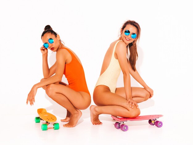 Twee mooie sexy glimlachende vrouwen in badpakken van de zomer de kleurrijke badmode. Trendy meisjes in zonnebril. Positieve modellen die op de vloer met kleurrijke stuiverskateboards zitten. geïsoleerd