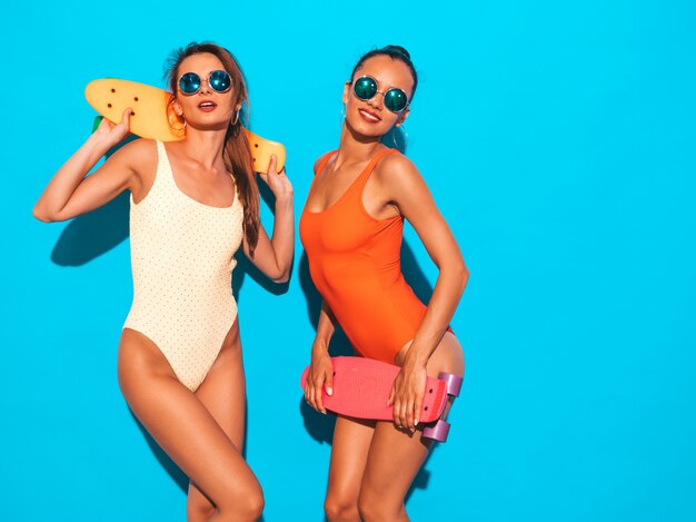 Twee mooie sexy glimlachende vrouwen in badpakken van de zomer de kleurrijke badmode. Trendy meisjes in zonnebril. Positieve modellen met plezier met kleurrijke penny skateboards. geïsoleerd